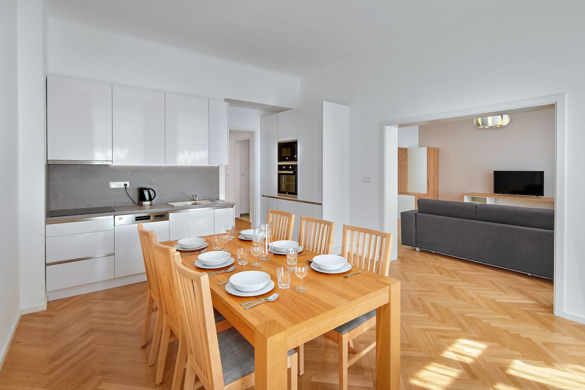 Uma cozinha moderna com armários brancos, uma mesa de jantar posta com pratos, copos e talheres, e um sofá cinza na sala de estar.
