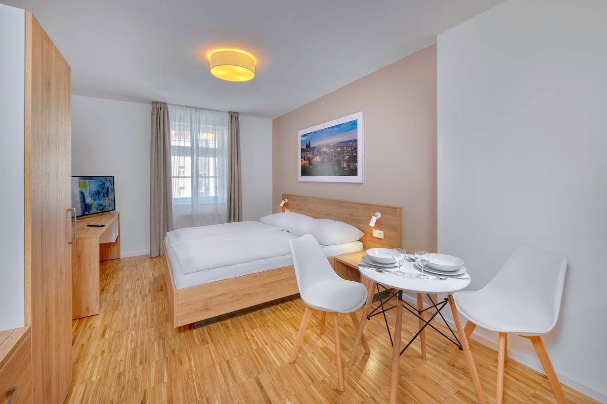 Quarto de hotel moderno com piso de madeira, cama com roupa de cama branca, escrivaninha com computador e área de refeições com duas cadeiras e uma mesa.