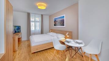 A modern hotel room with a wooden floor, a bed with white linens, a desk with a computer, and a dining area with two chairs and a table.