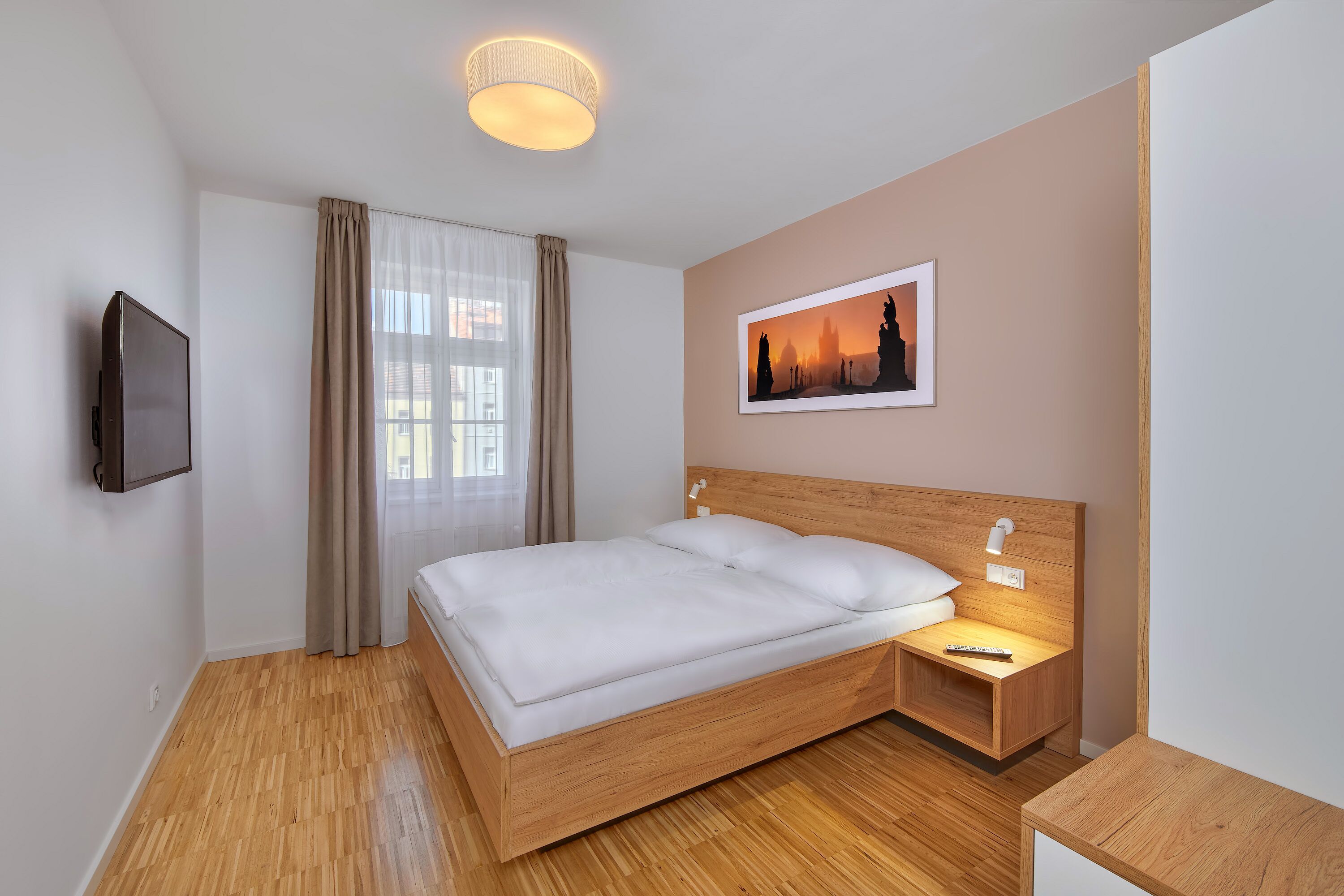 A neatly arranged hotel room with a wooden bed, a bedside table with a lamp, a flat-screen TV mounted on the wall, and a framed picture on the wall.