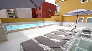 Un espace piscine avec des chaises longues et des parasols, une petite piscine et un bâtiment en arrière-plan.