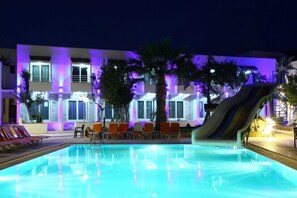 Una piscina con sillas de descanso y un tobogán, rodeada de palmeras y un edificio con iluminación púrpura.