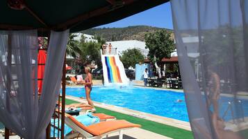 Un área de piscina con tobogán, sillas de descanso y vista a una colina.