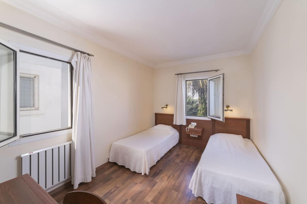 A small, well-lit room with two single beds, a wooden floor, a window with curtains, a radiator, and a small table.
