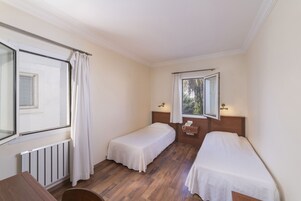 Una habitación pequeña y luminosa con dos camas individuales, suelo de madera, una ventana con cortinas, un radiador y una mesita.