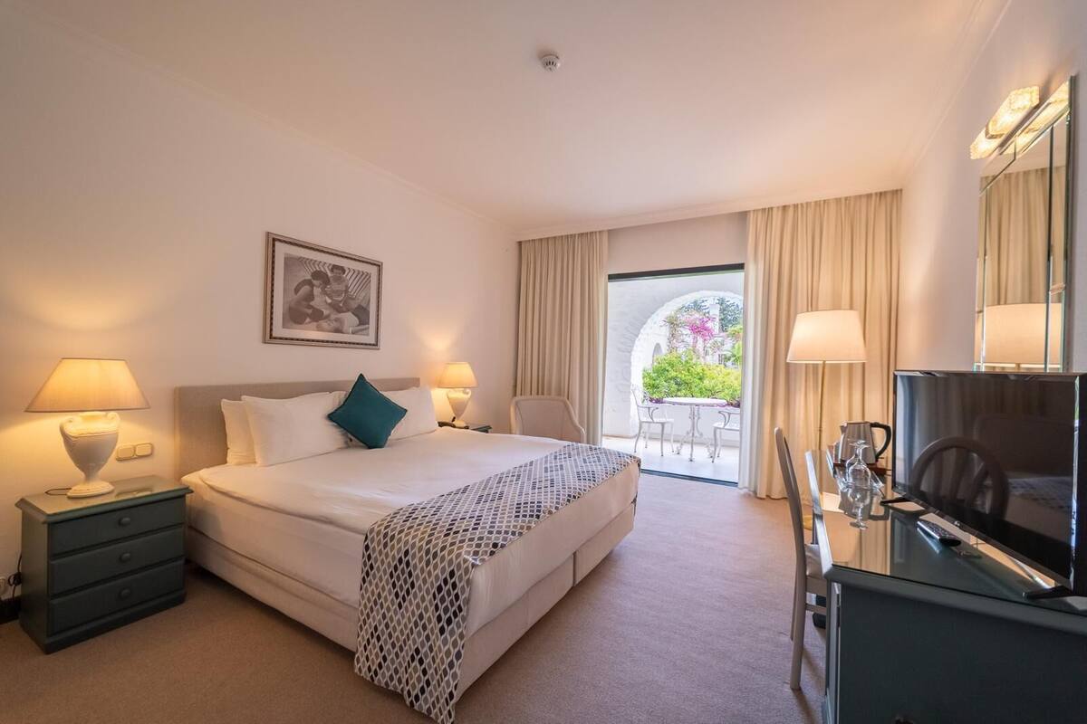 Ein Hotelzimmer mit Bett, Nachttischen, einem Schreibtisch mit Fernseher und einem Balkon mit Vorhängen.