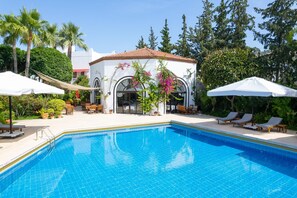 Piscina con sillas de descanso y sombrillas, rodeada de vegetación y un edificio con ventanas arqueadas.