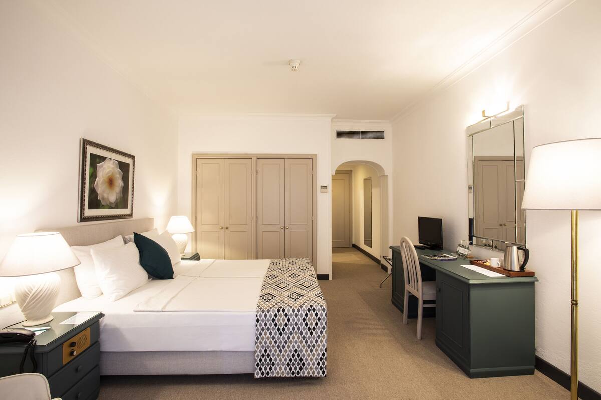 Ein Hotelzimmer mit Bett, Nachttischlampen, einem Schreibtisch mit Stuhl, einem Spiegel und einem Kleiderschrank.