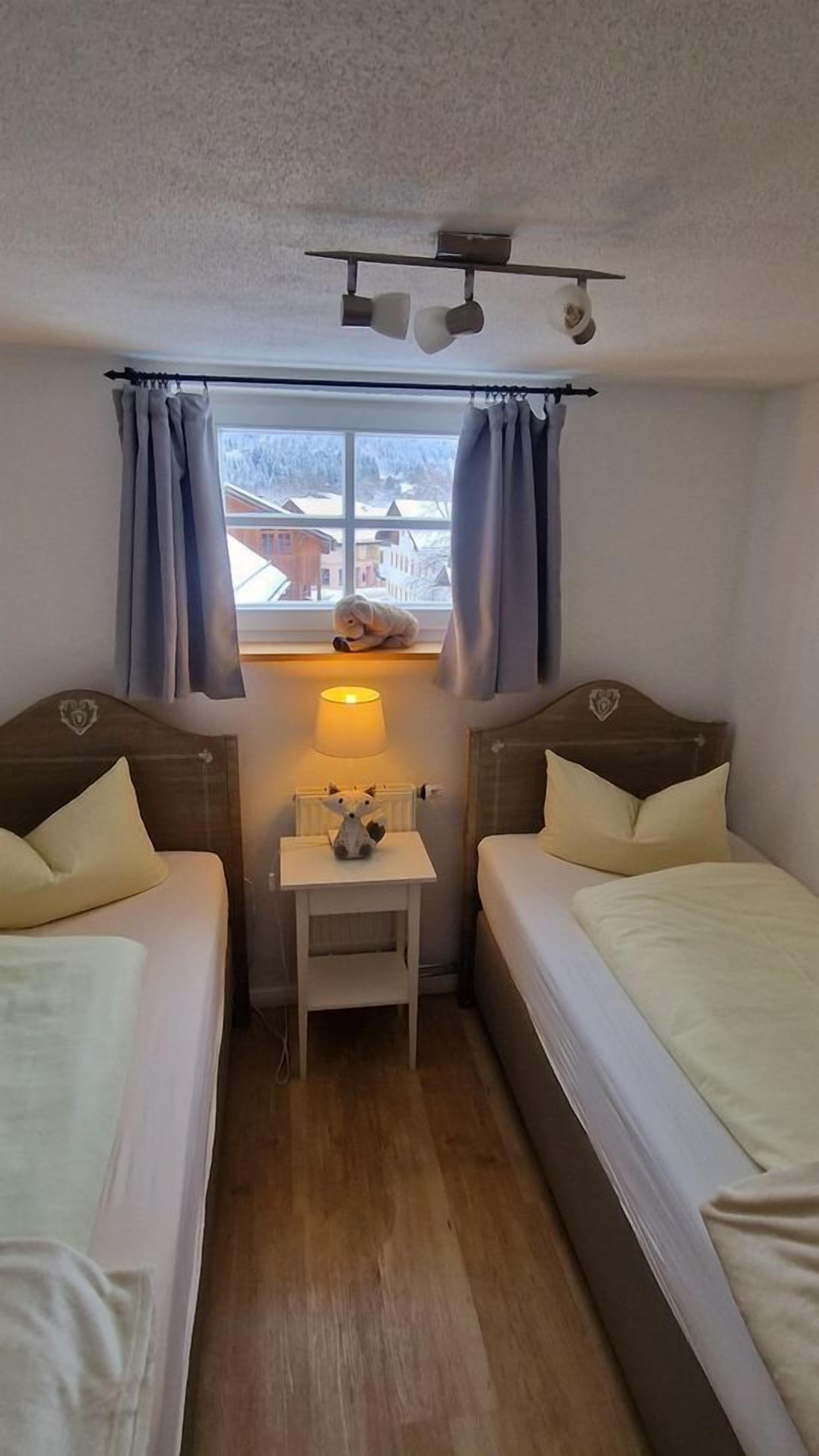 A small, cozy room with two beds, a nightstand with a lamp, and a window with curtains.