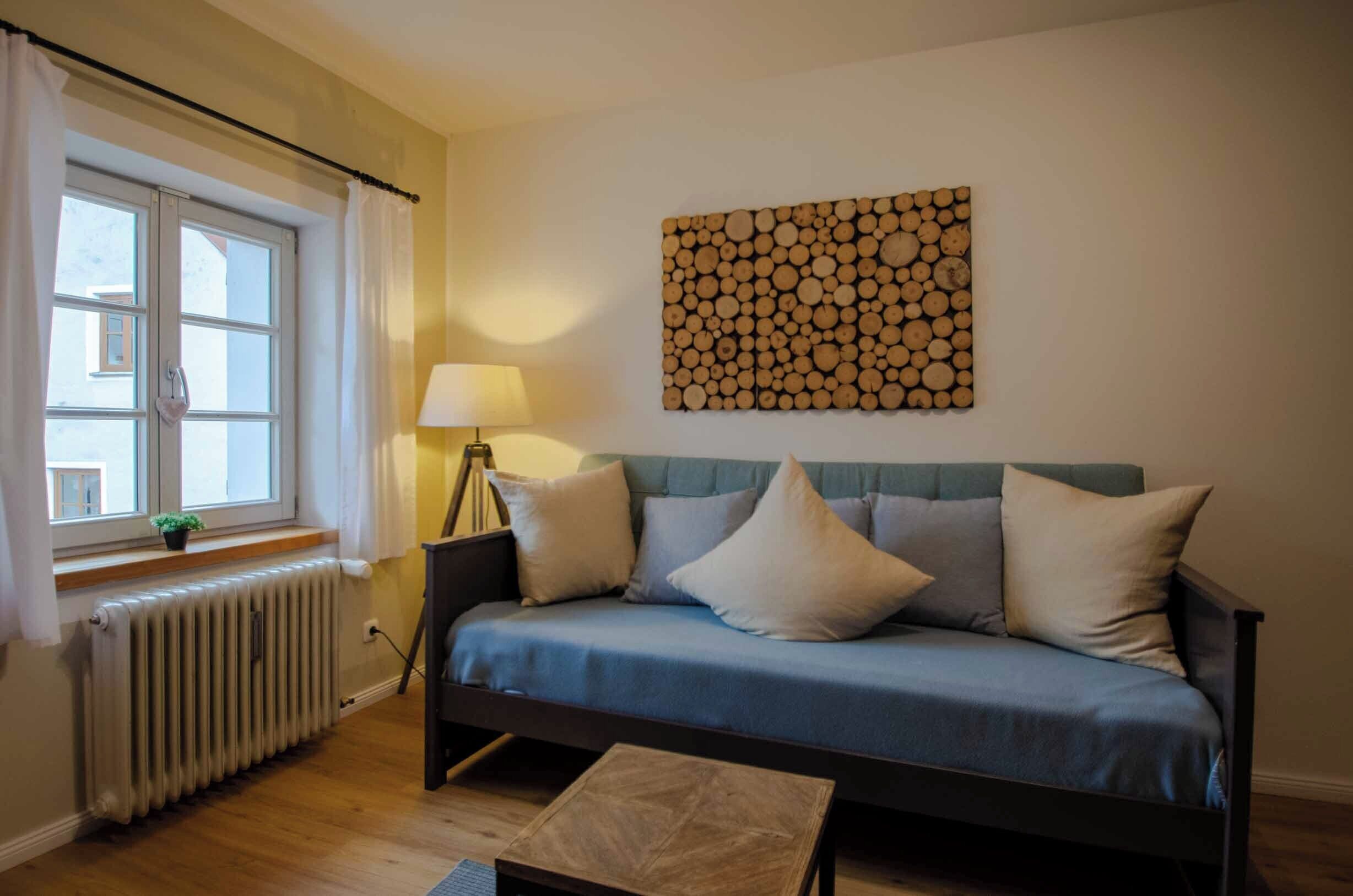 A cozy room with a blue sofa, a wooden log wall art, a window with curtains, a radiator, and a lamp.
