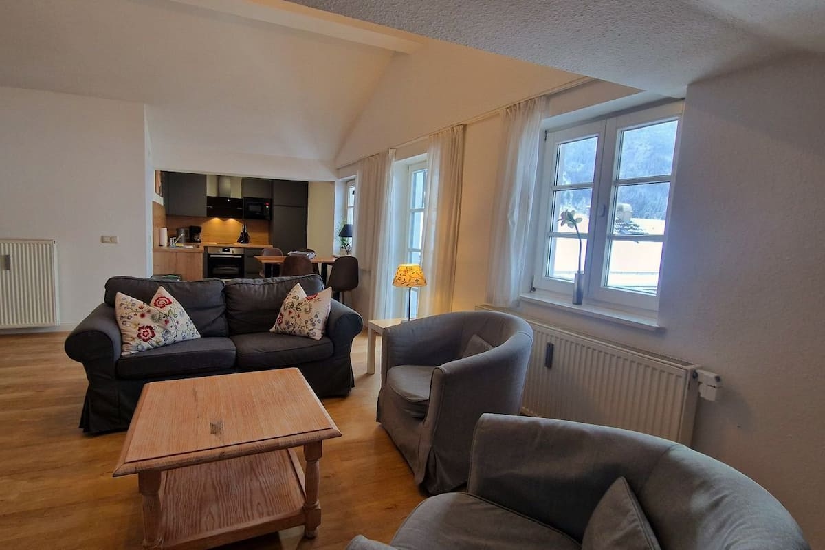 A spacious living area with a sofa set, a wooden coffee table, and a grey armchair. A kitchen is visible in the background.