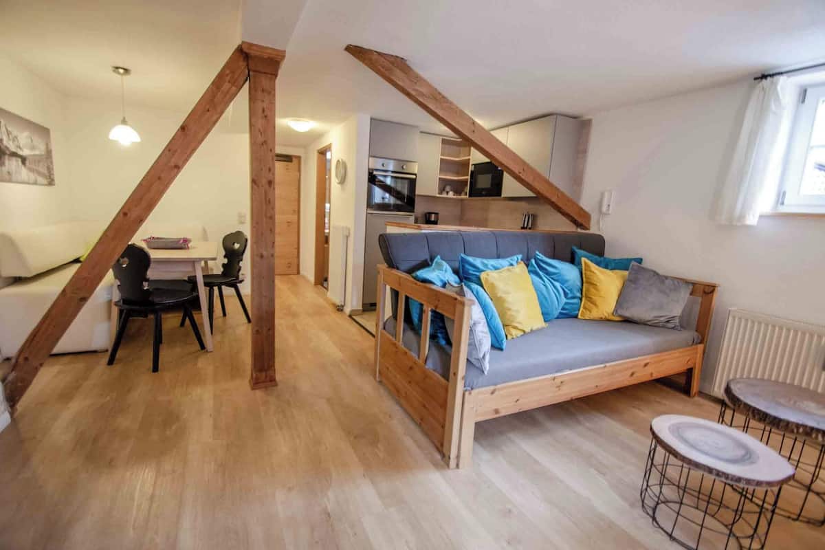 A cozy living room with a wooden ceiling, a sofa with cushions, a dining table with chairs, and a kitchen area in the background.