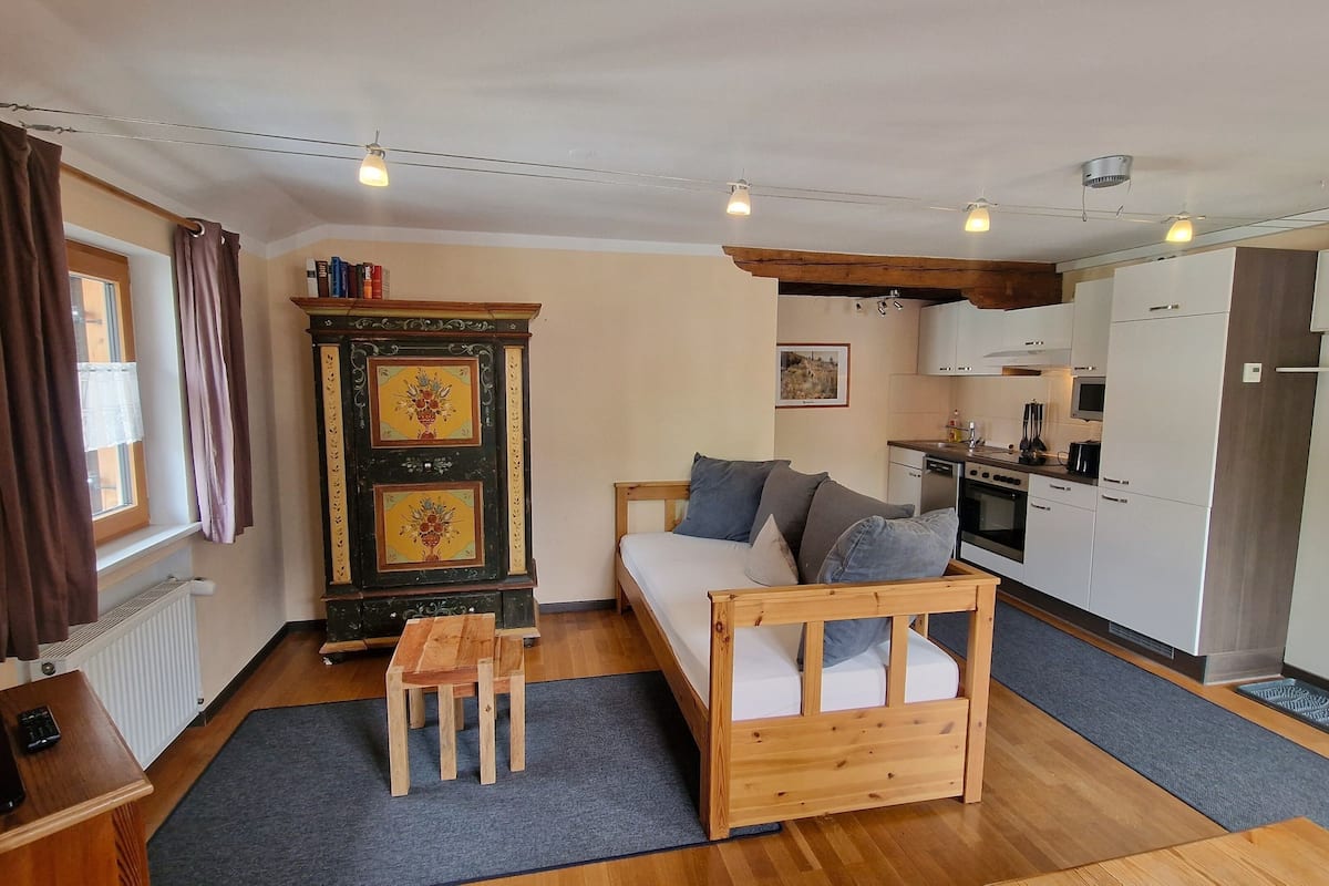 A compact living space with a bed, a small wooden table, a kitchenette, and a cabinet with decorative panels.