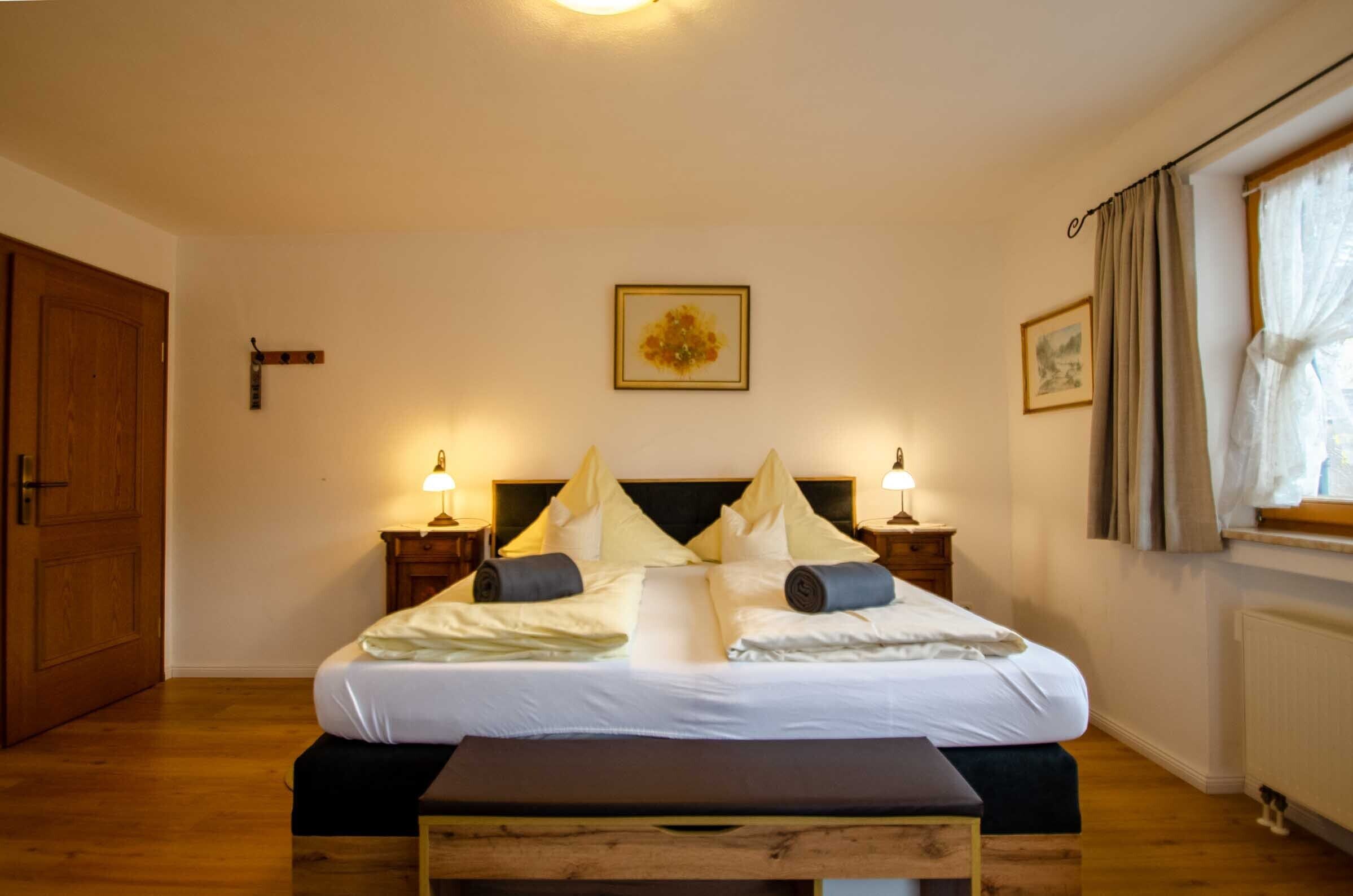 A neatly made bed with pillows and a blanket, wooden headboard, bedside tables with lamps, a wooden bench, and a window with curtains.