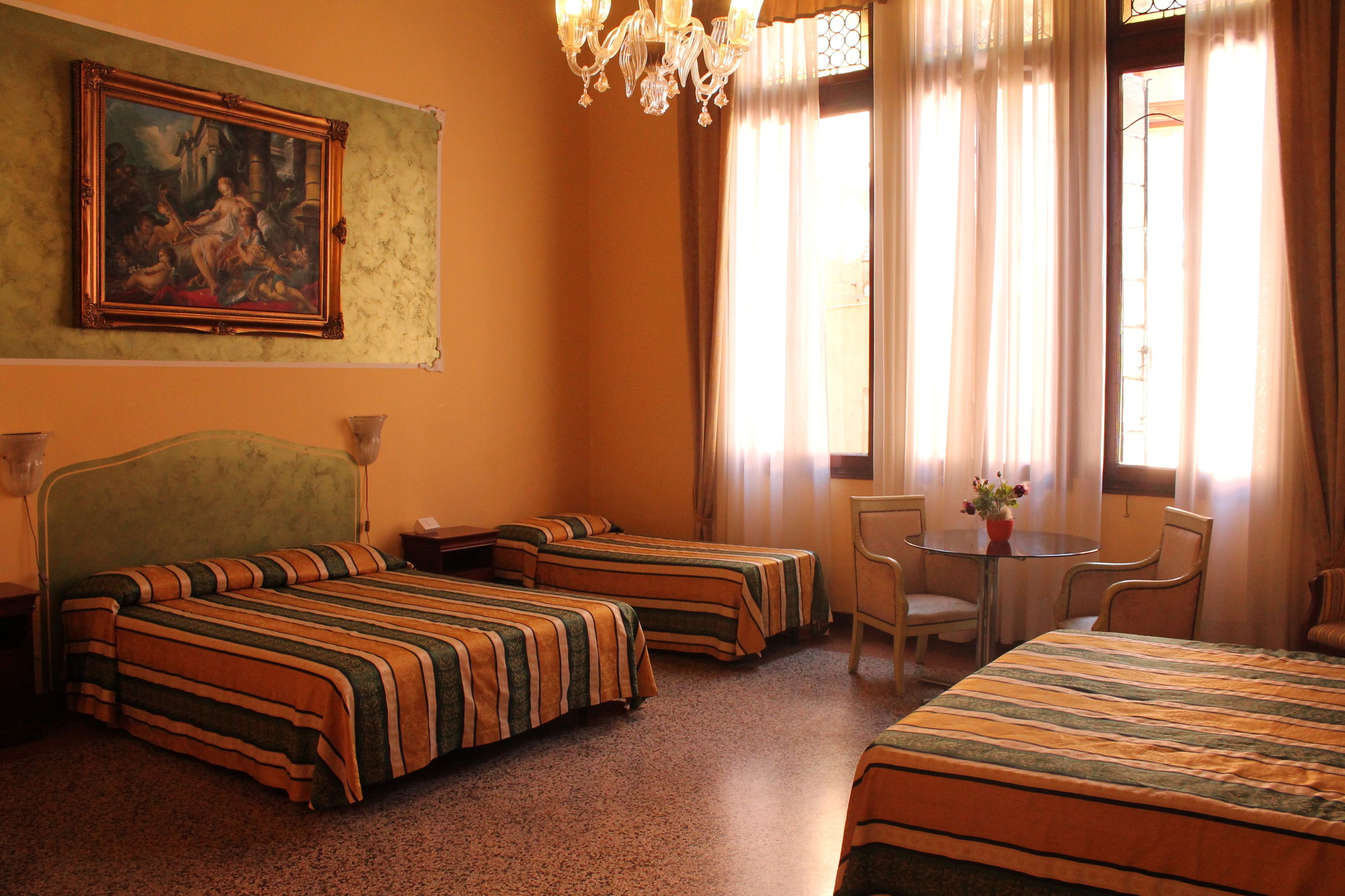 A hotel room with two single beds, a dining table, chairs, a chandelier, and a painting on the wall.
