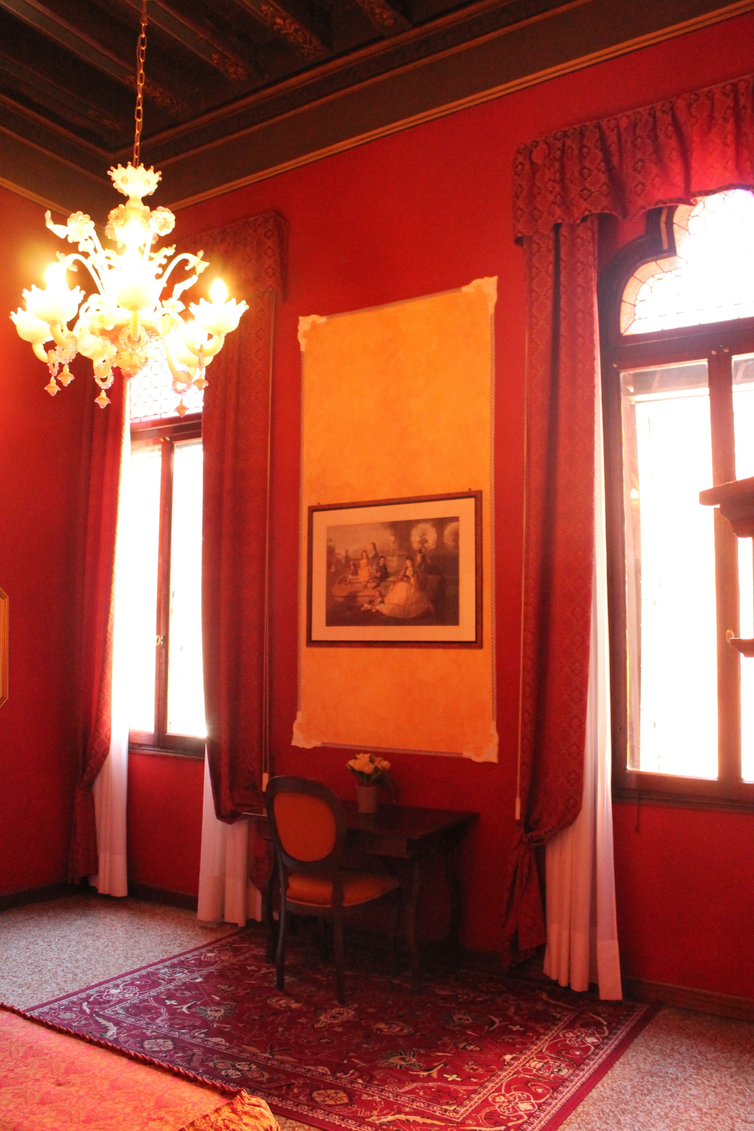 A room with red walls, a chandelier, a framed picture, a table with chairs, and a window with curtains.
