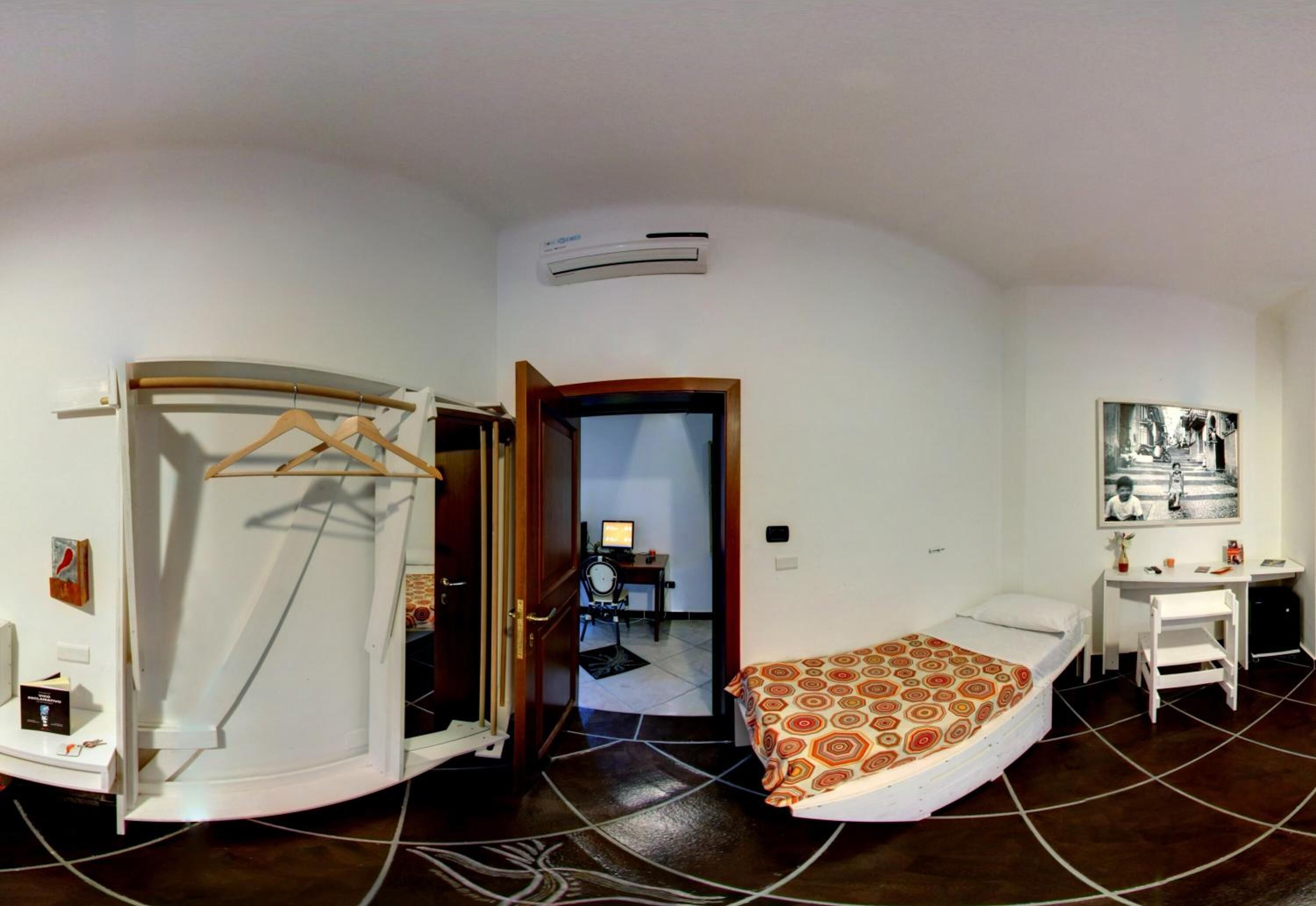 Una stanza compatta con un letto, una scrivania e una sedia. Una porta dà accesso a un'altra stanza con una scrivania e una sedia. Un condizionatore d'aria è montato a parete.
