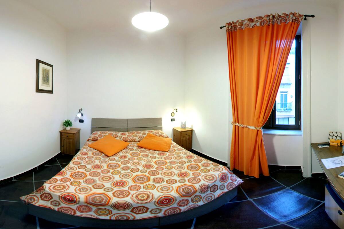 Ein Schlafzimmer mit einem Bett, einem Nachttisch, einer Lampe und einem Fenster mit Vorhängen.