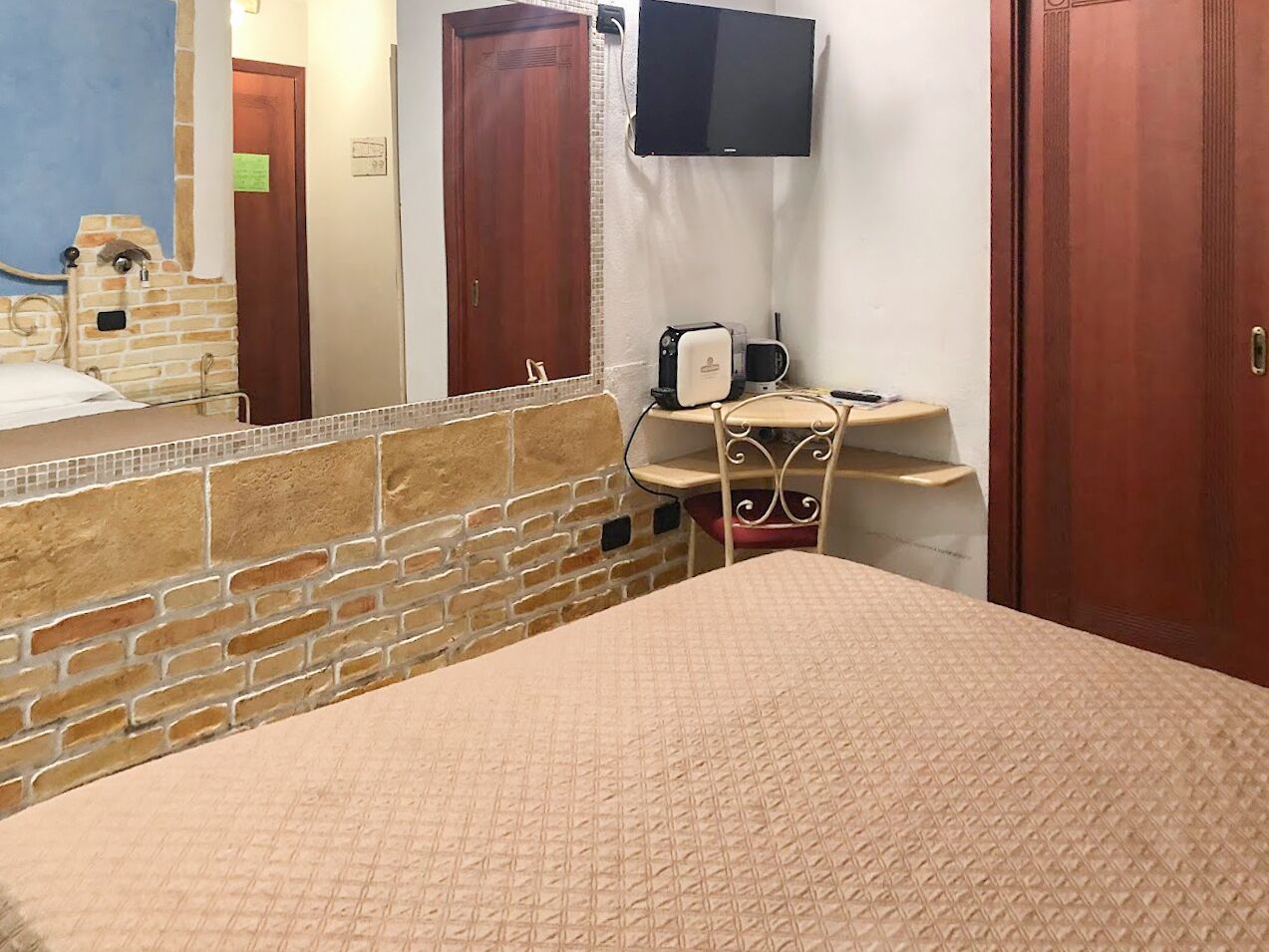 A hotel room with a brick wall, a bed with a beige quilt, a small desk with a chair, a television, and a wooden door.