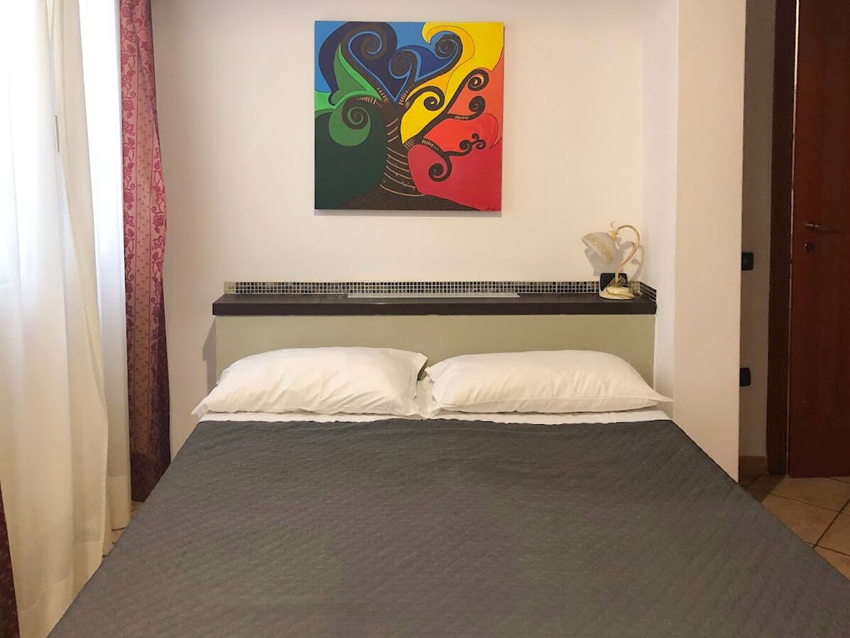 Een bed met een grijze spreidek en witte kussens, daarboven een kleurrijk abstract schilderij, en een klein tafeltje met een lamp en een banaan erop.