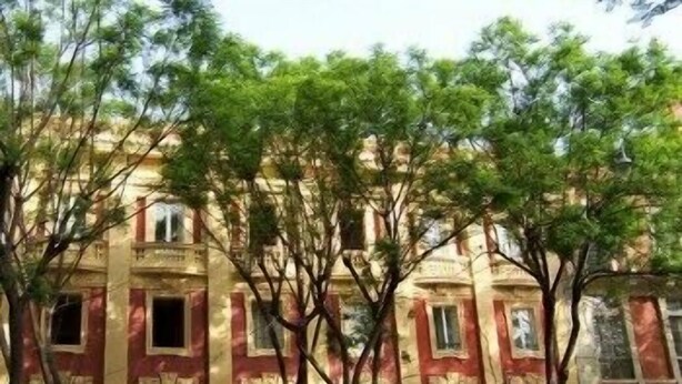A building with a red and beige facade, multiple windows, and trees in front. - Sardinia Domus (Cagliari)