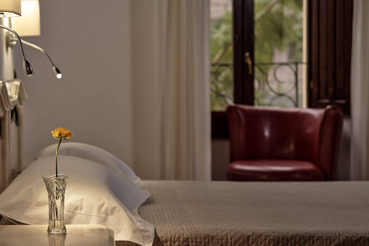 A bed with a white pillow, a vase with a single yellow flower, and a red leather chair in a hotel room.