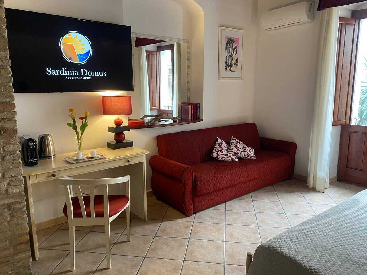 Una habitación con un sofá rojo, un escritorio con lámpara, un televisor en la pared y una ventana con cortinas.