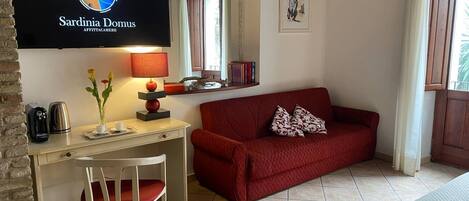 Una habitación con un sofá rojo, un escritorio con lámpara, un televisor en la pared y una ventana con cortinas.