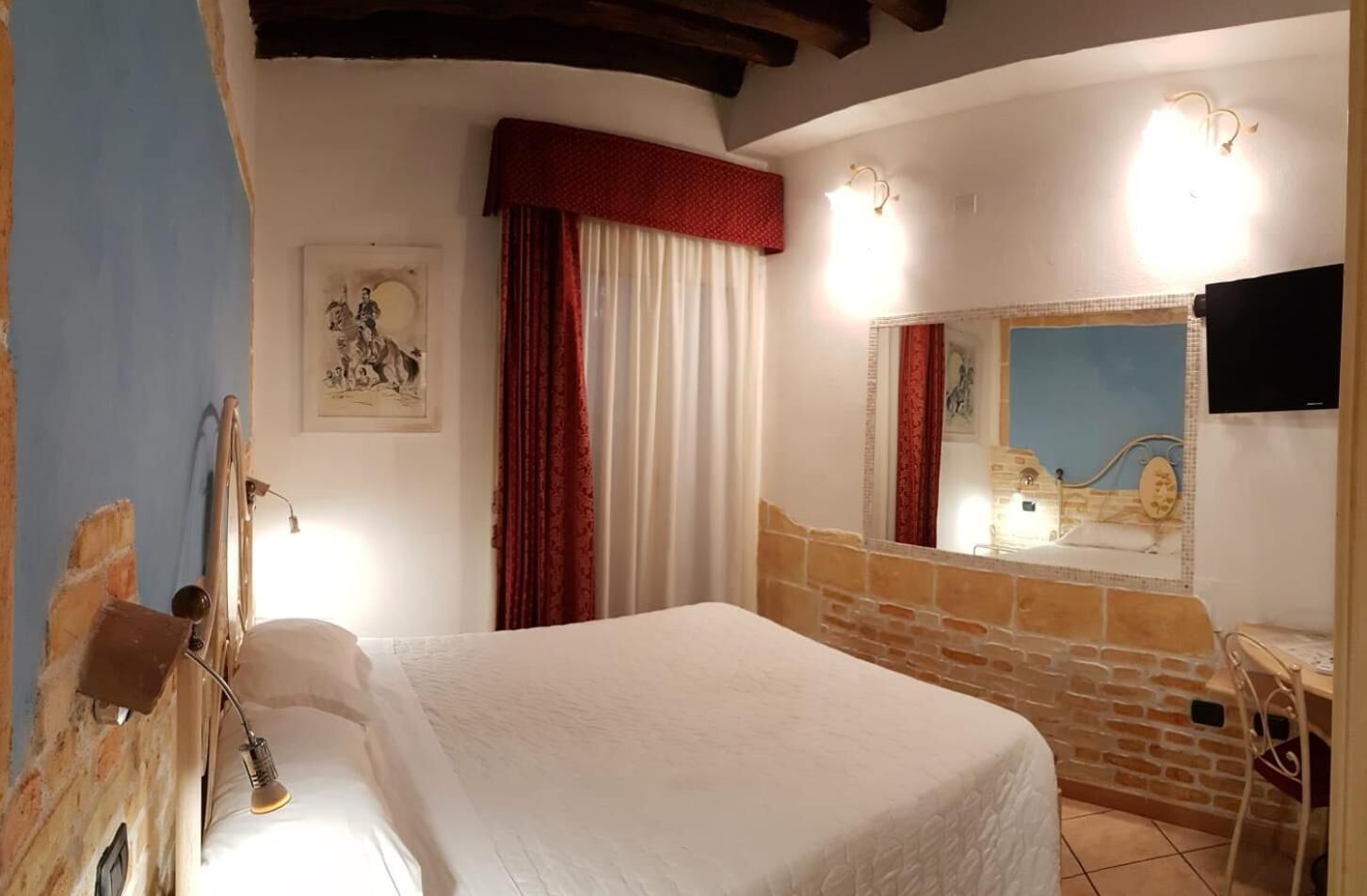 A bedroom with a bed, a mirror, a wall-mounted TV, and a window with red curtains.