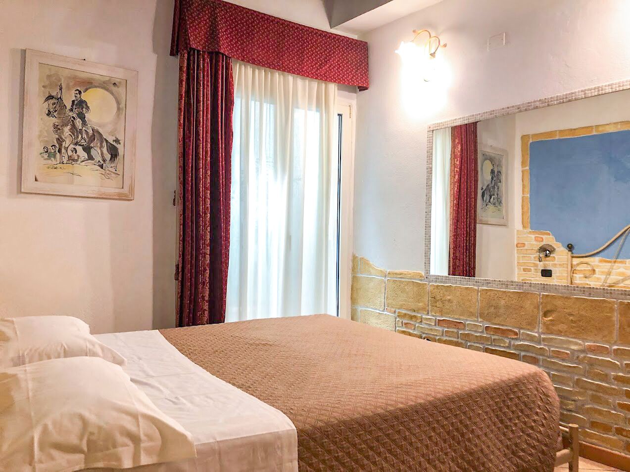 A bedroom with a bed, a large window with curtains, a stone wall, and a mirror.
