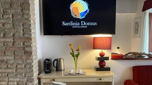 A room with a brick wall, a desk with a chair, a television displaying 'Sardinia Domus', a lamp, and a red chair. - Sardinia Domus (Cagliari)