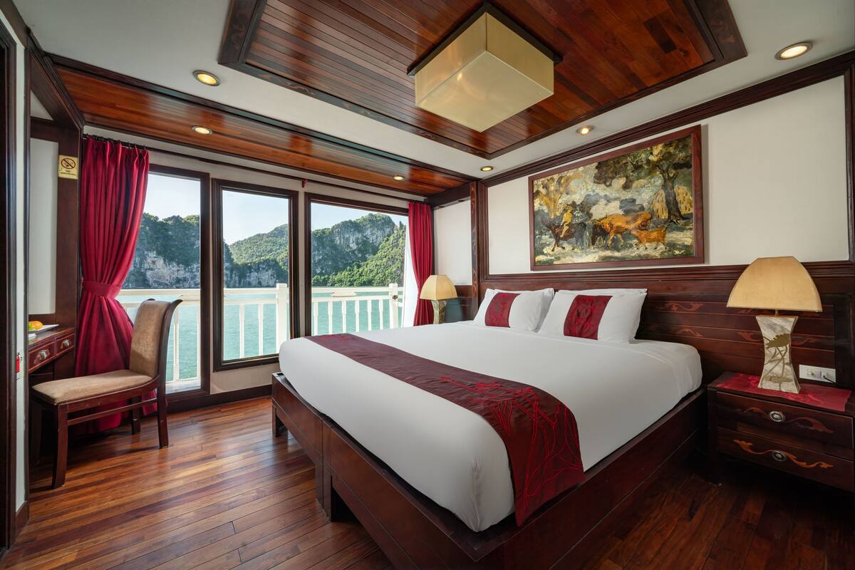 A large bed with a red and white bedspread, wooden flooring, a painting on the wall, and a view of the outdoors through the windows.
