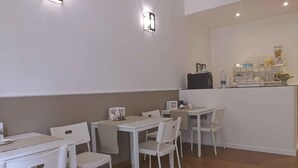 A dining area with white tables and chairs, a counter with various items, and wall-mounted lighting. - Possidonea 28 Bed & Breakfast (Reggio Calabria)
