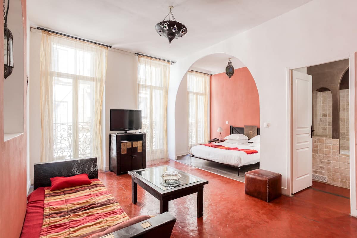 A hotel room with a bed, a sofa, a television, a glass coffee table, and a bathroom visible through an archway.