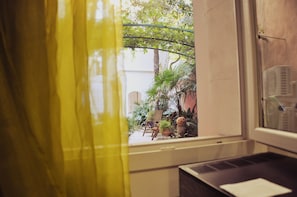 Blick aus einem Fenster mit gelbem Vorhang, der einen kleinen Außenbereich mit einem Stuhl, Topfpflanzen und einer Treppe zu einer oberen Ebene zeigt.