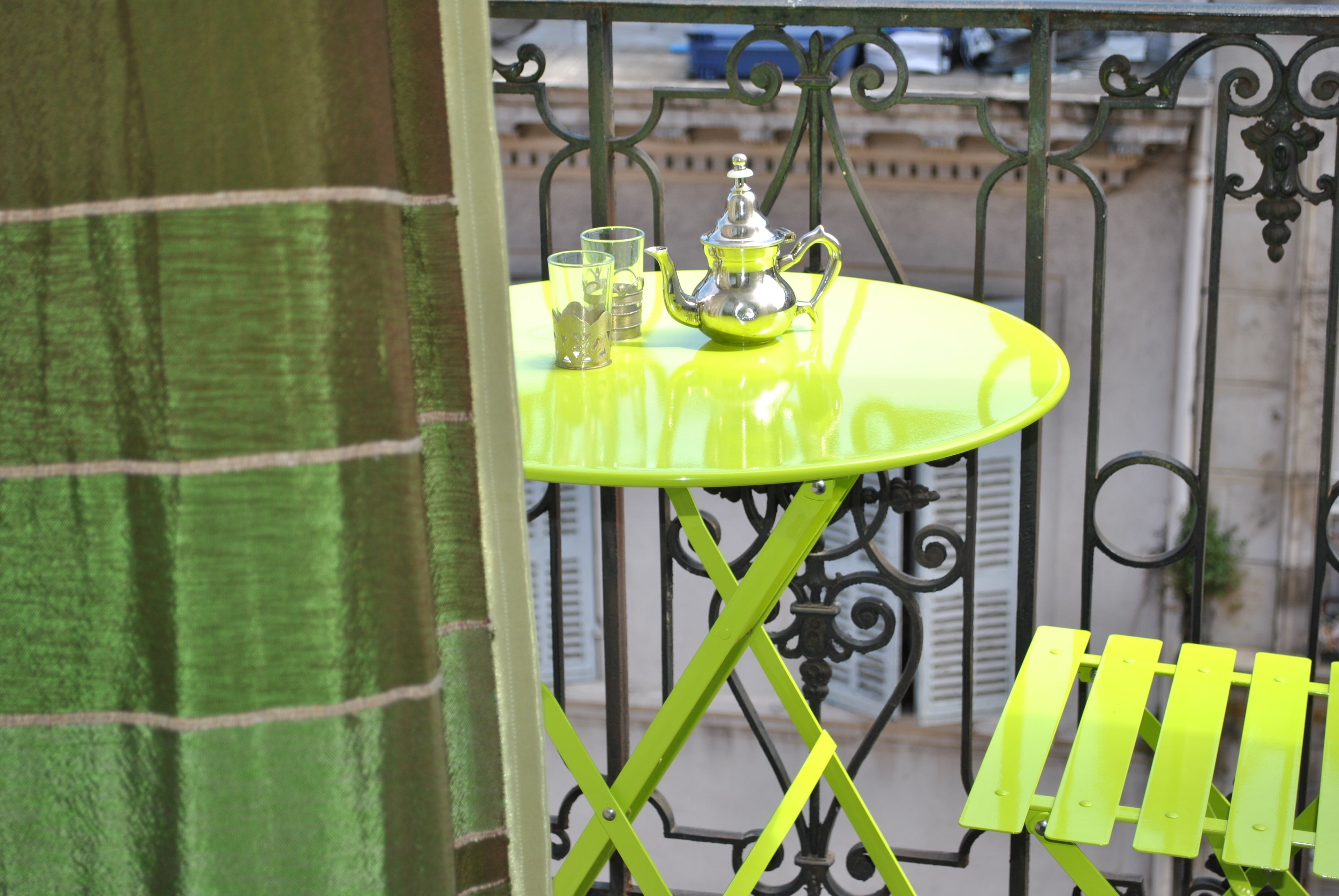 Een kleine ronde tafel met een groen metalen frame en een felgele blad. Op de tafel staan een zilveren theepot, twee glaasjes en een kleine schaal.