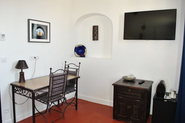 A room with a dining table, chairs, a television, a cabinet, and a wall-mounted shelf with a decorative plate.