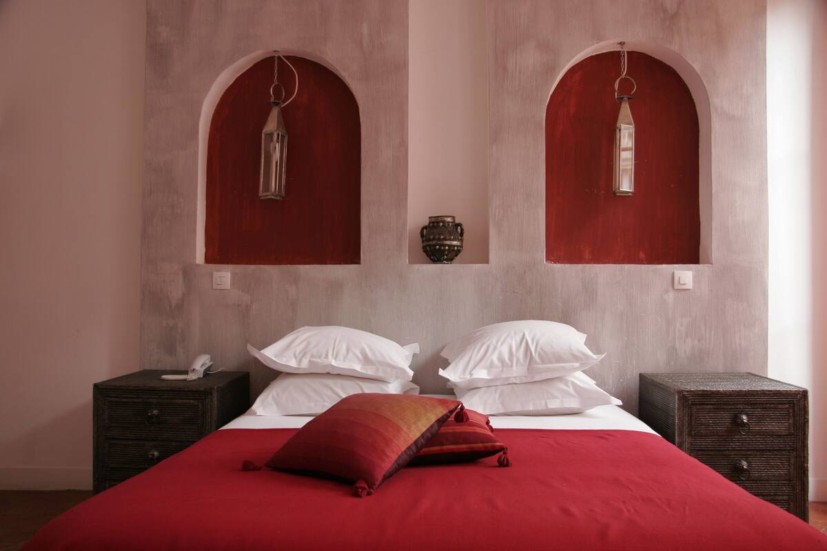 A bedroom with a large bed, two bedside tables, a red bedspread, white pillows, and two wall-mounted lanterns.