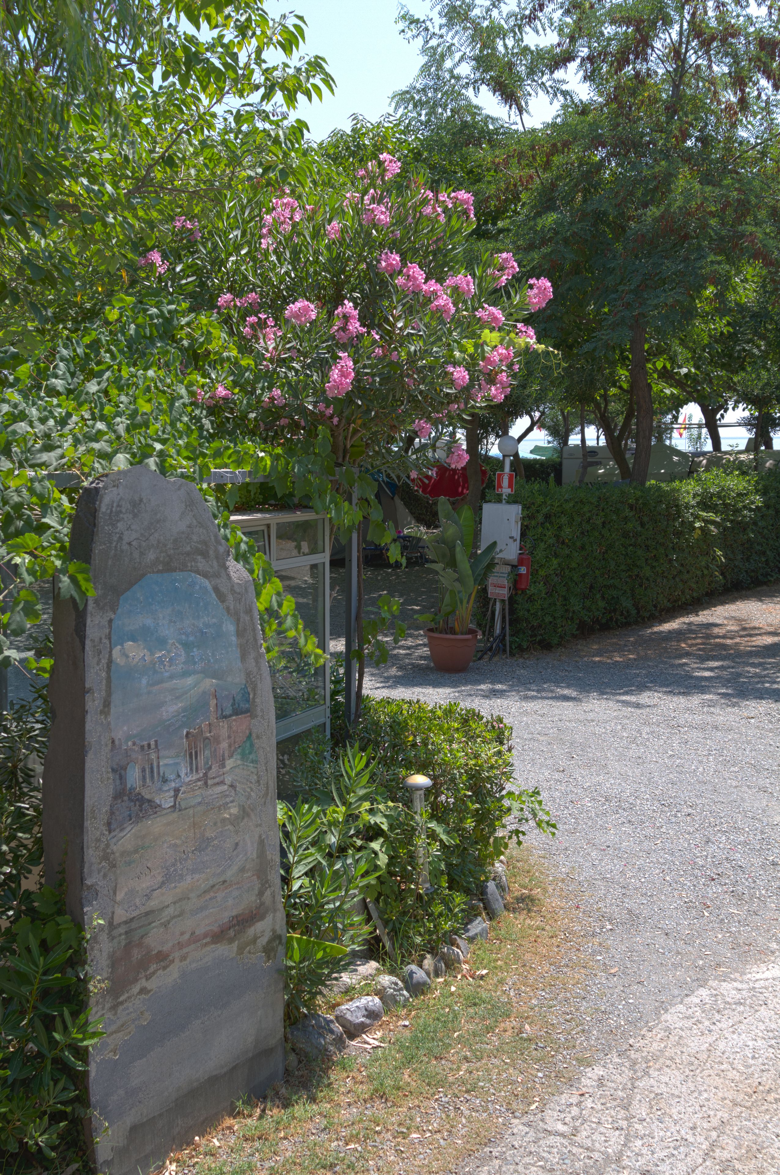 A stone sign with a painted landscape, a flowering tree, and a potted plant beside a gravel path.