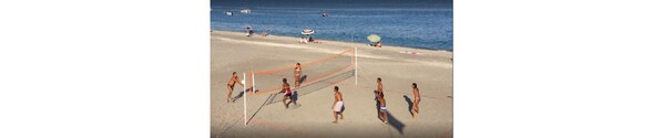 Partido de voleibol de playa con jugadores en traje de baño, una red y gente en el fondo.