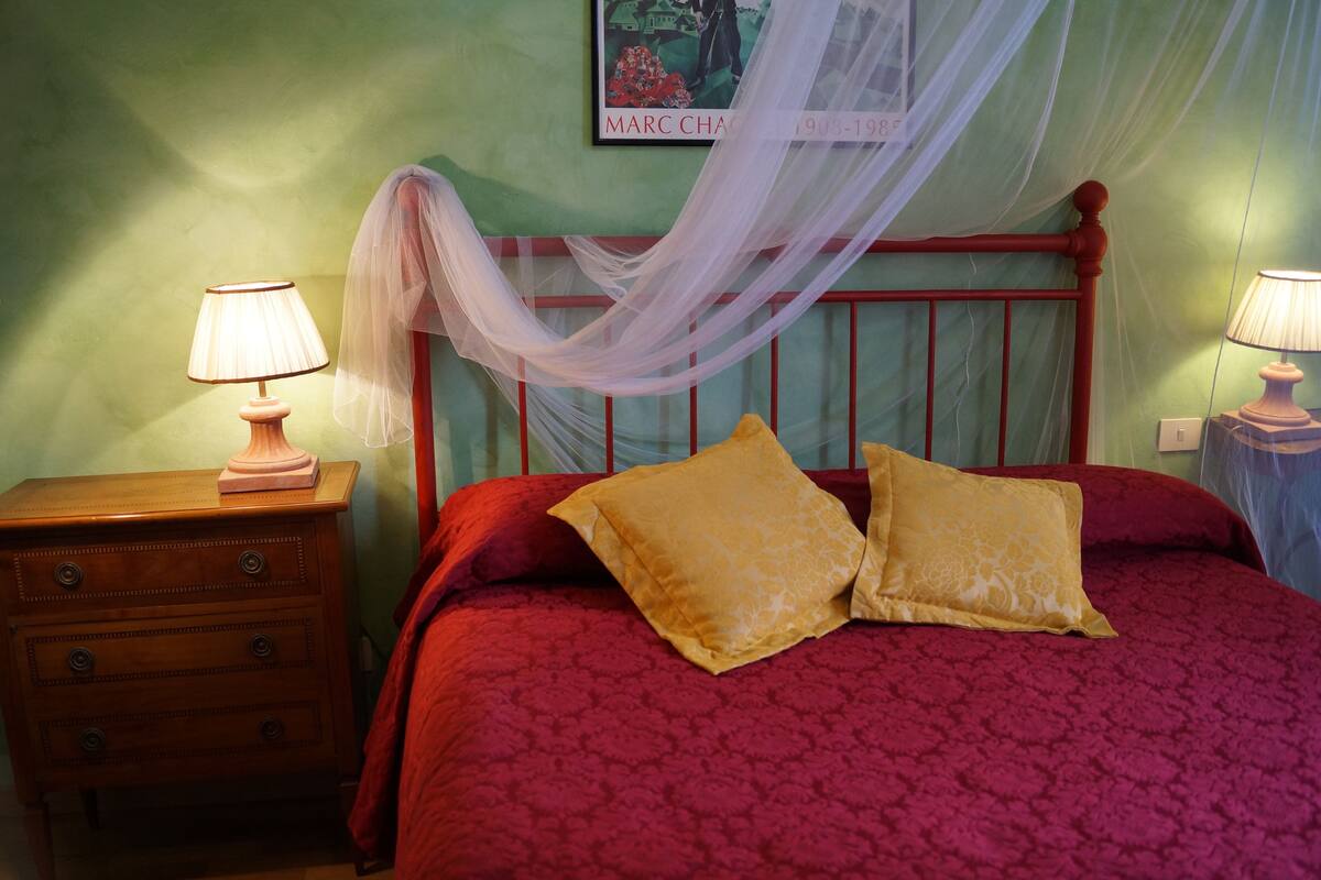 A bed with a red headboard, two yellow pillows, and a red bedspread. There is a wooden nightstand with a lamp on it to the left.