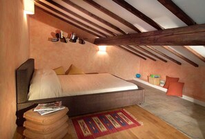 Quarto com cama, mesinha, cadeira e estante com livros e objetos decorativos.