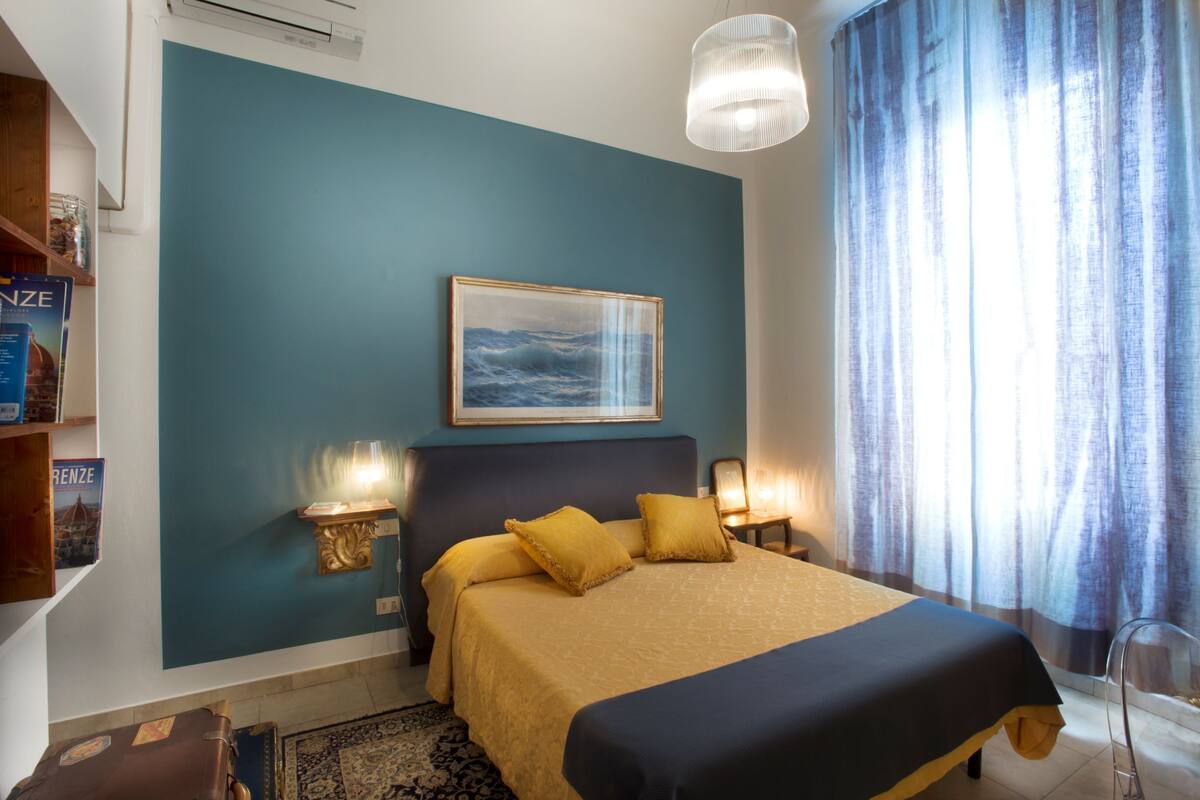 A bedroom with a bed, a painting, a nightstand, a lamp, and a suitcase.