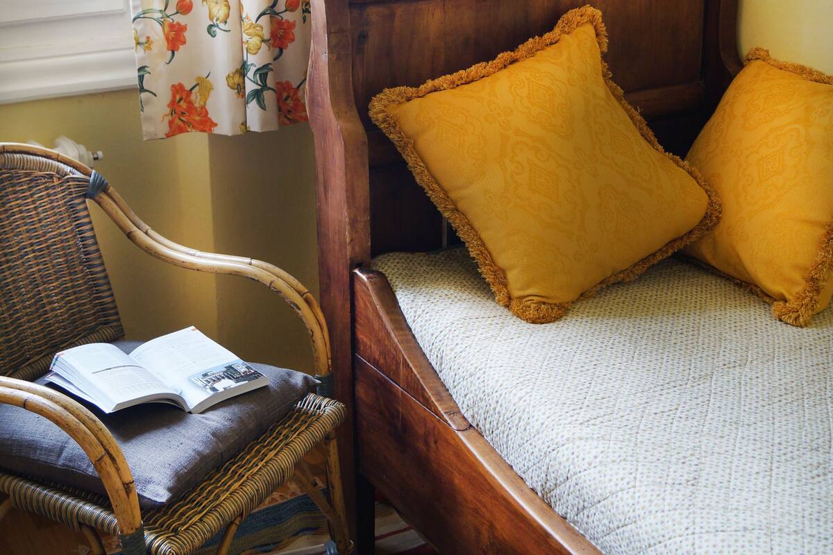 A wooden bed with yellow pillows, a wicker chair, and a floral curtain.