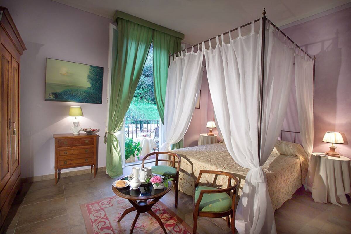 A bedroom with a four-poster bed, a nightstand with a lamp, a chair, and a view of the outdoors through a window with green curtains.