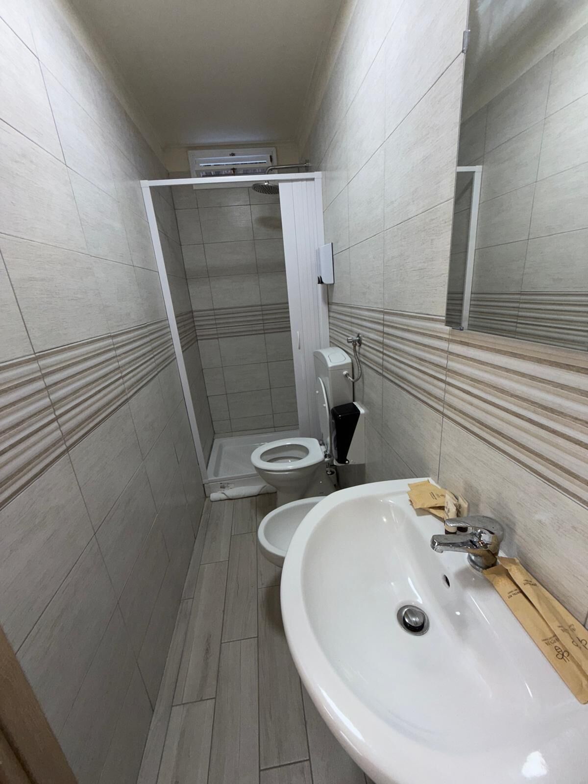 A narrow bathroom with a white sink, a toilet, and a shower area.
