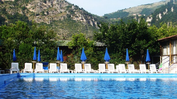 A swimming pool with lounge chairs and blue umbrellas, surrounded by greenery and mountains.