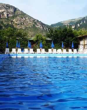 Une piscine entourée de verdure et de montagnes, avec des chaises longues et des parasols bleus.