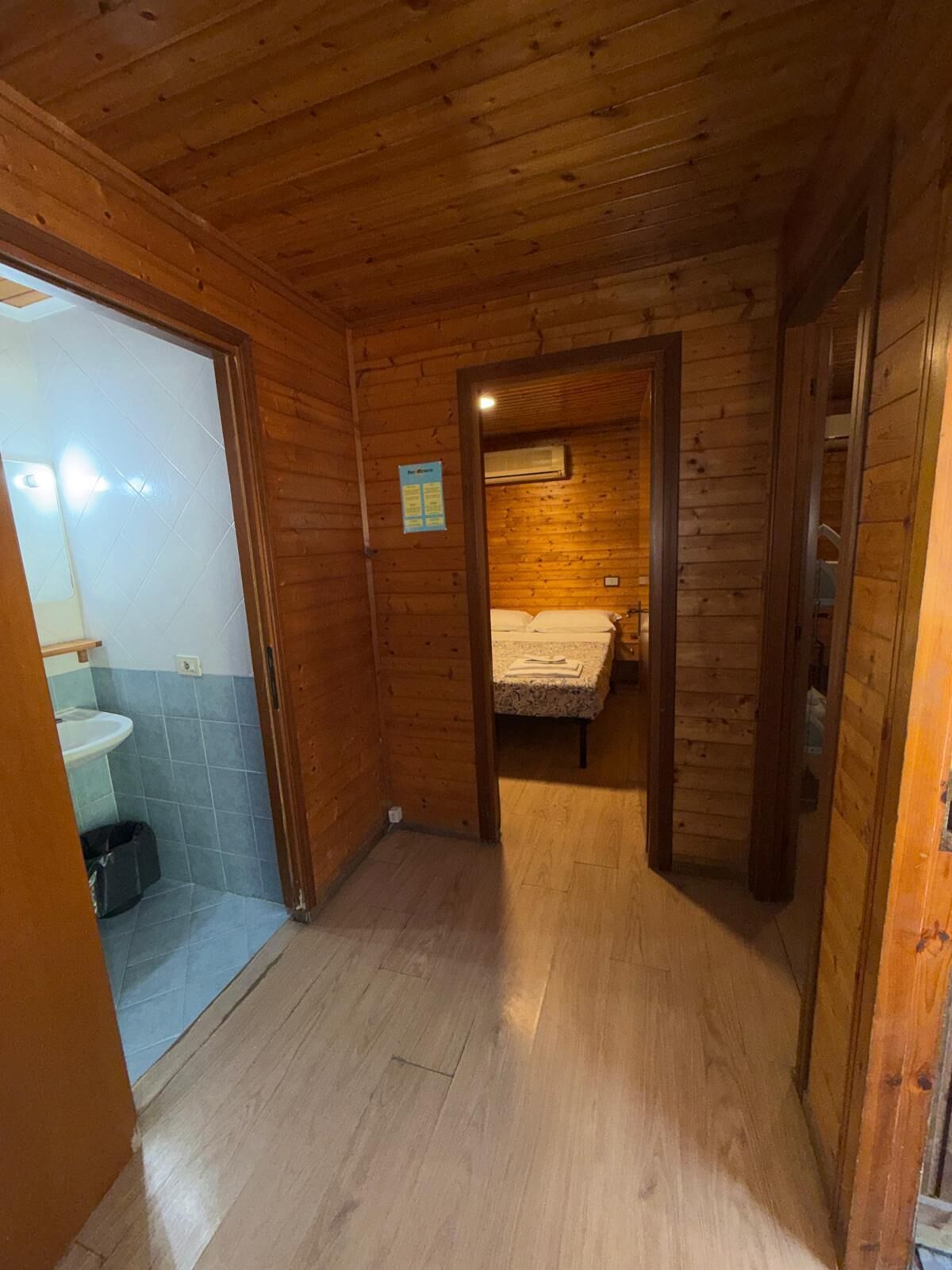Interior de madera con un baño a la izquierda, una cama con manta en el centro y una pequeña mesa con un libro.