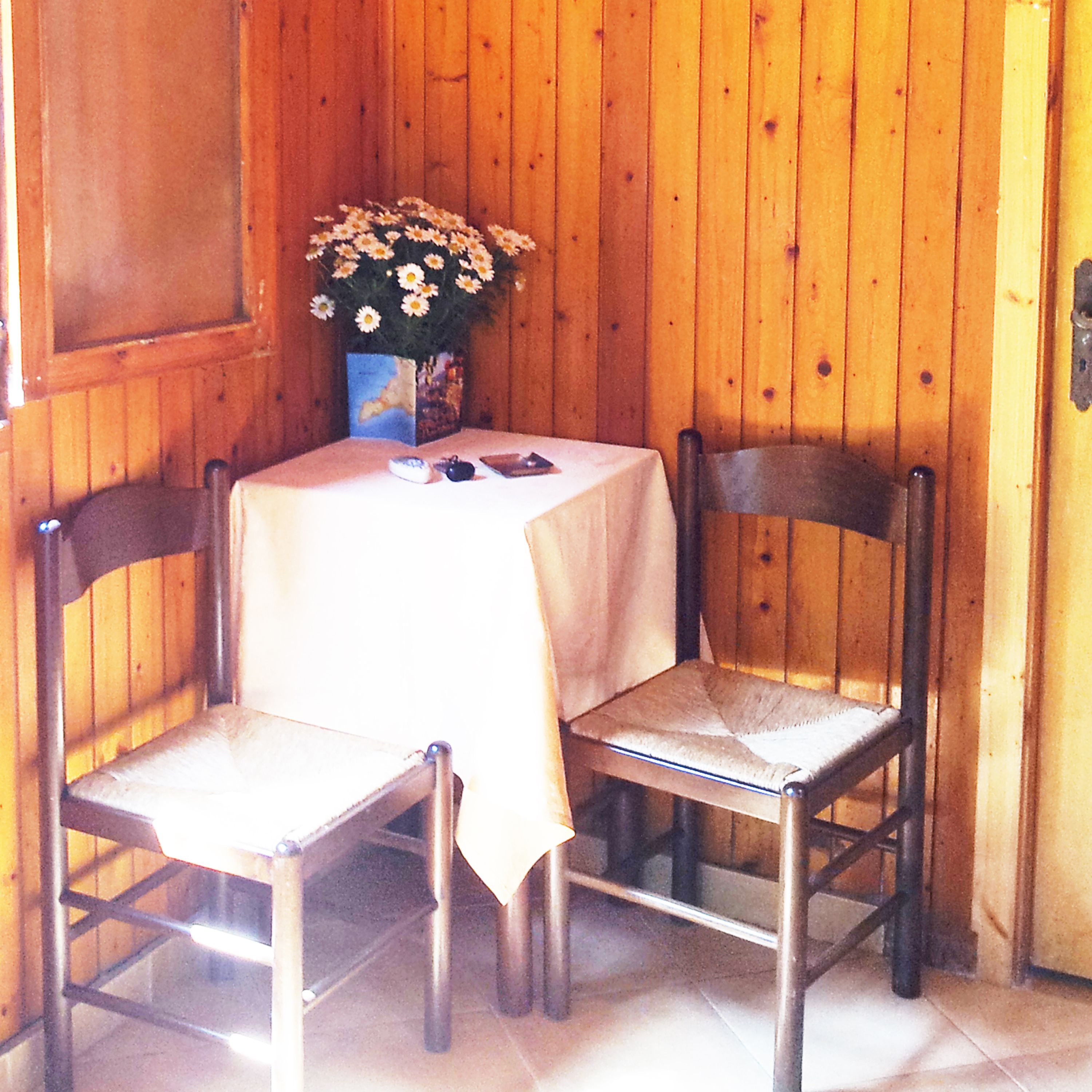 A wooden interior with a table and two chairs, a vase with flowers, and a painting on the wall.